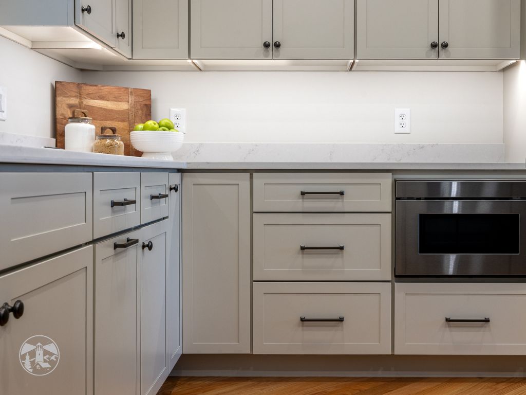 Modern kitchen renovation by Lighthouse Contracting Group in New Hampshire’s Lakes Region with gray shaker cabinets, under-cabinet lighting, quartz countertops, and built-in stainless wall oven.